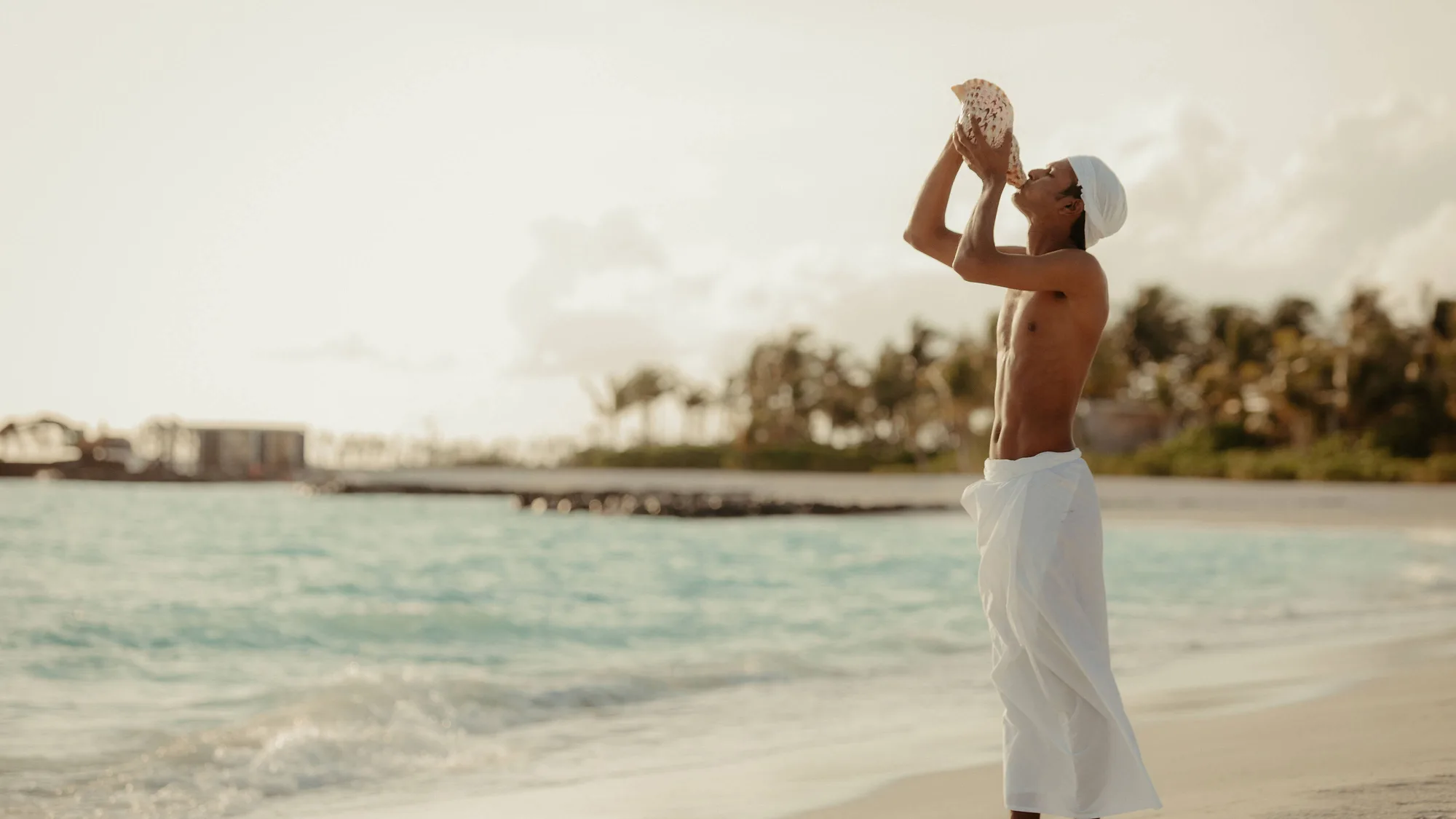 A resort host blowing a conch shell on the beach at a traditional welcome ceremony